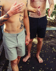 Two shirtless barefoot men standing next to each other in a parking lot. One is wearing solid gray boardshorts and the other is wearing solid black boardshorts.