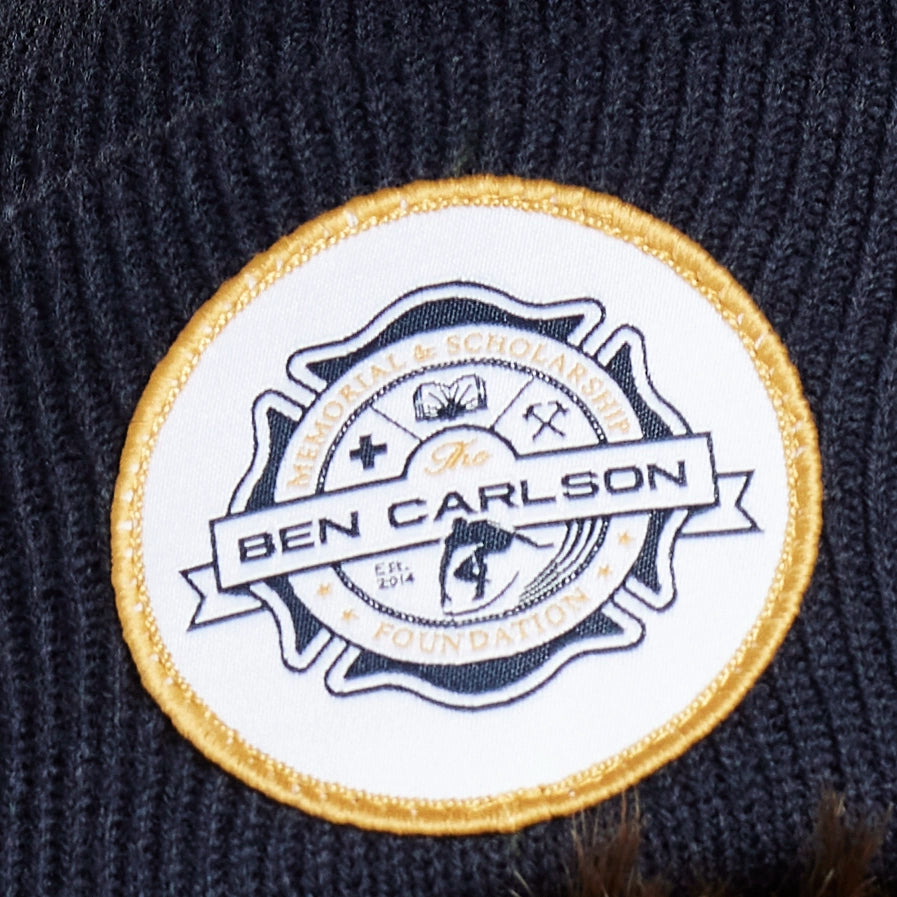 White circular patch with gold trim and a fire department-style badge in the center. Across the badge on a banner are the words The Ben Carlson. Arching above the banner are the words Memorial and Scholarship. Arching below the banner is the word Foundation with two stars on each side. There's a small image of a surfer crouching and riding a wave on his surfboard below the banner.
