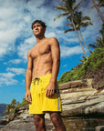 A shirtless man wearing yellow boardshorts standing on the edge of a rocky tropical coastline.