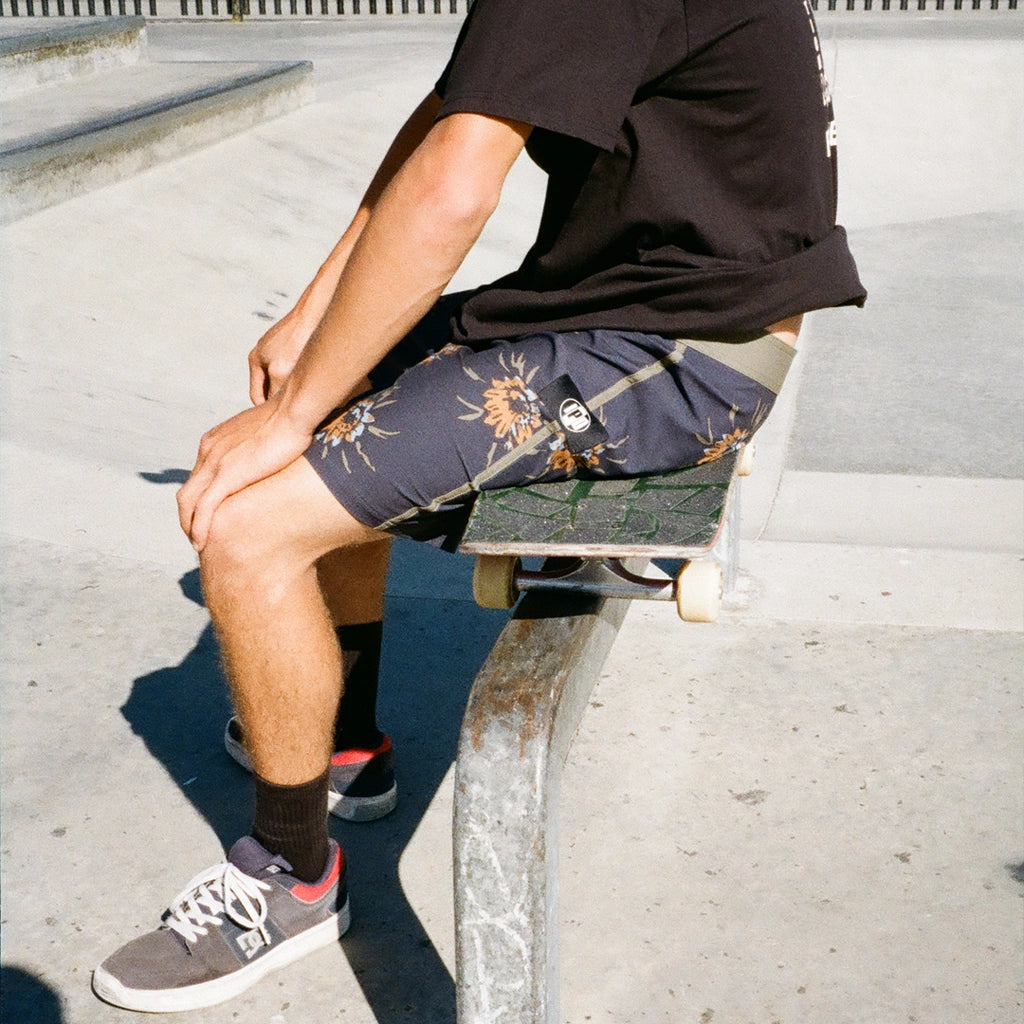A skater sits on his skateboard while wearing black boardshorts with a floral pattern.