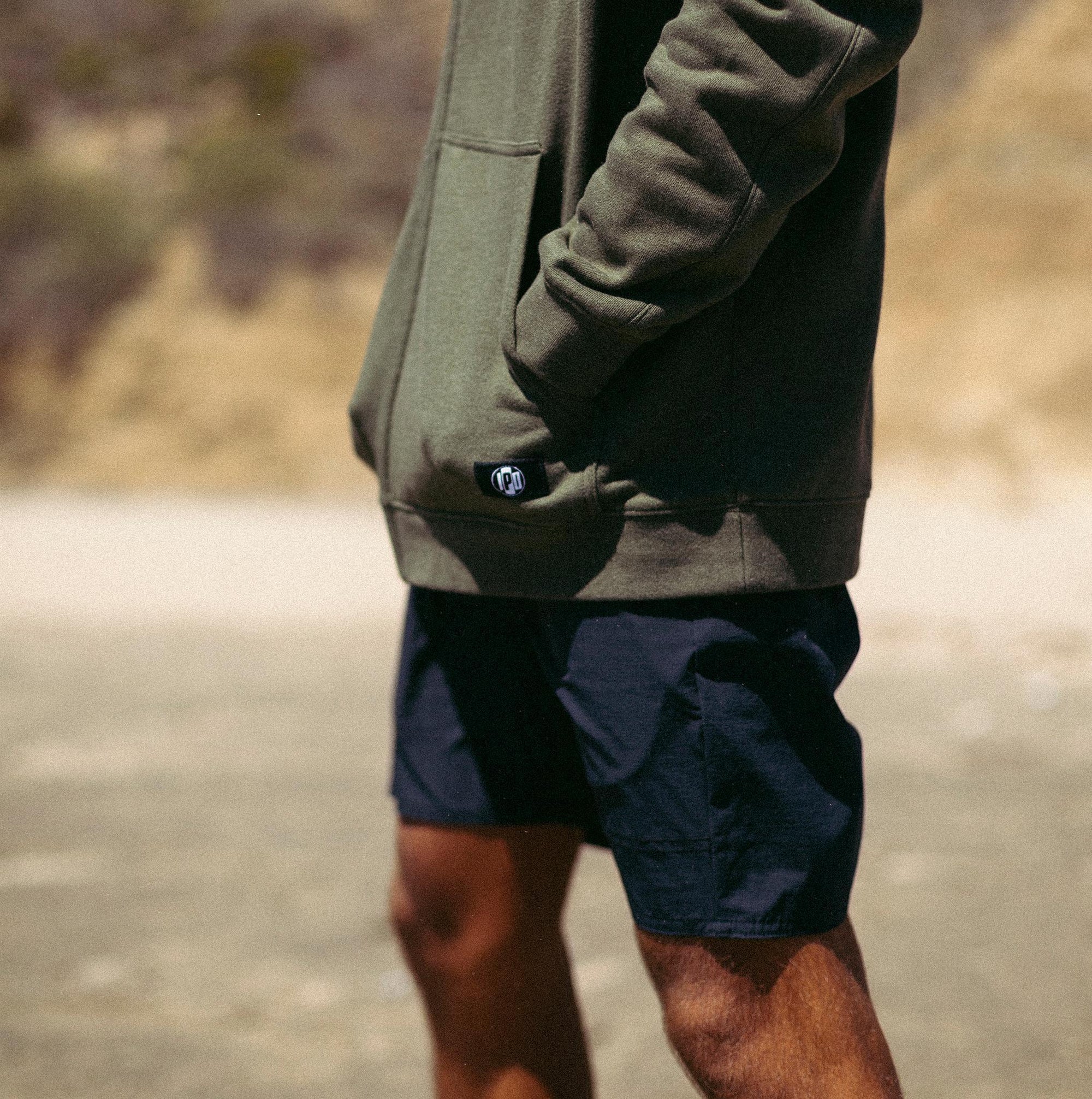 Side view of a man standing outside wearing an olive green hoodie and solid black lightweight boardshorts.